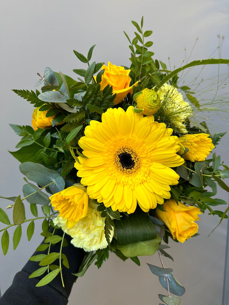 Kukkakimppu, jossa on keltainen gerbera, sekä keltaista jaloleinikkiä, neilikkaa ja ruusua.
